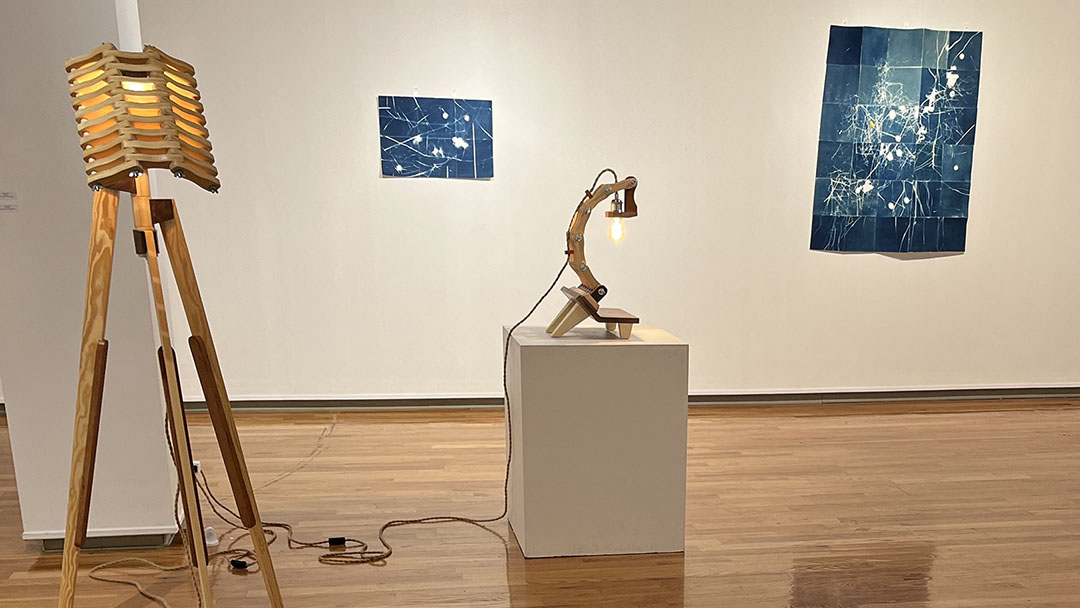 two wooden sculptures in front of two cyanotypes in a gallery with a hardwood floor and a white wall